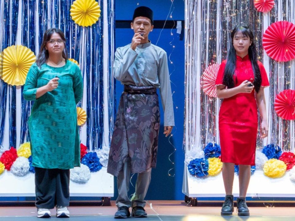 Three student emcees, elegantly dressed in traditional attire, lead the festive Hari Raya assembly.